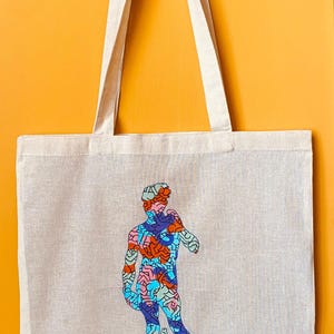 May include: Beige tote bag with a colorful graphic of the David statue. The artwork features a mosaic-like design with blue, orange, and red hues. The bag has long, beige handles and a small signature in the bottom left corner.
