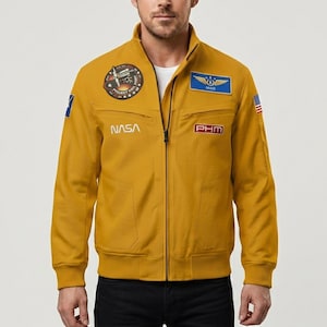 May include: Mustard yellow zip-up jacket with NASA and other patches. The jacket has a stand-up collar, two front pockets, and a small American flag patch on the right sleeve. The jacket has the text "PROJECT MAIL MARY" on a circular patch.