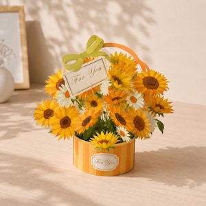May include: A paper bouquet of sunflowers and daisies in a striped container. The bouquet has a green bow and a tag that says "For You". The container is orange and yellow. A charming gift for any occasion.