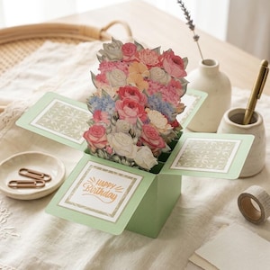 May include: A light green pop-up birthday card with a 3D floral arrangement in pink, white, and blue. The card has a gold-coloured "Happy Birthday" message. The card is open, revealing the paper flowers. Other items on the table include a pen and washi tape.