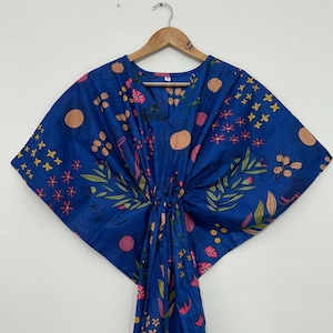 May include: A blue kaftan featuring a vibrant botanical print with pink, yellow, and peach-colored flowers, leaves, and circular shapes. The garment has a V-neckline, wide sleeves, and a drawstring waist detail, displayed on a wooden hanger.