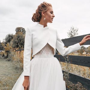 May include: A white bridal gown featuring a long, flowing tulle skirt and a cropped, long-sleeved jacket. The jacket has a high collar and is paired with a lace bodice. The bride is holding a small bouquet of greenery.