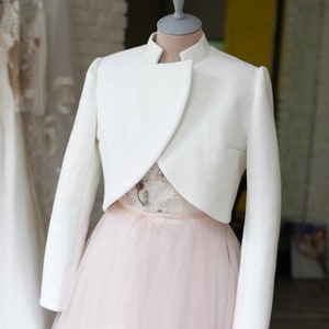 May include: A white, long-sleeved bolero jacket with a high collar, displayed on a mannequin. The jacket is paired with a light pink tulle skirt and a detailed bodice. Other white dresses hang in the background.