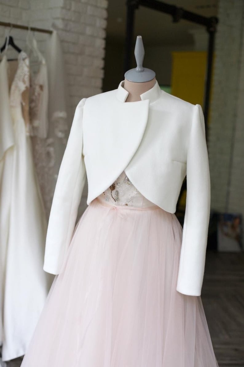 May include: A white, long-sleeved bolero jacket with a high collar, displayed on a mannequin. The jacket is paired with a light pink tulle skirt and a detailed bodice. Other white dresses hang in the background.