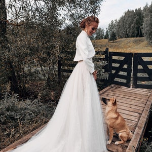 May include: A bride in a white wedding dress with a long train stands on a wooden bridge. A small dog sits on the bridge, looking up at the bride. The scene is set outdoors with trees and a fence in the background.