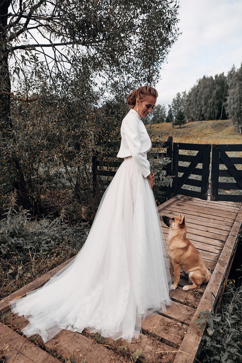 May include: A bride in a white wedding dress with a long train stands on a wooden bridge. A small dog sits on the bridge, looking up at the bride. The scene is set outdoors with trees and a fence in the background.