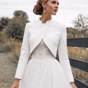May include: A white bridal bolero jacket with long sleeves and a high collar, paired with a white tulle skirt. The jacket is tailored and covers the shoulders, creating an elegant silhouette. The background features a blurred outdoor scene.