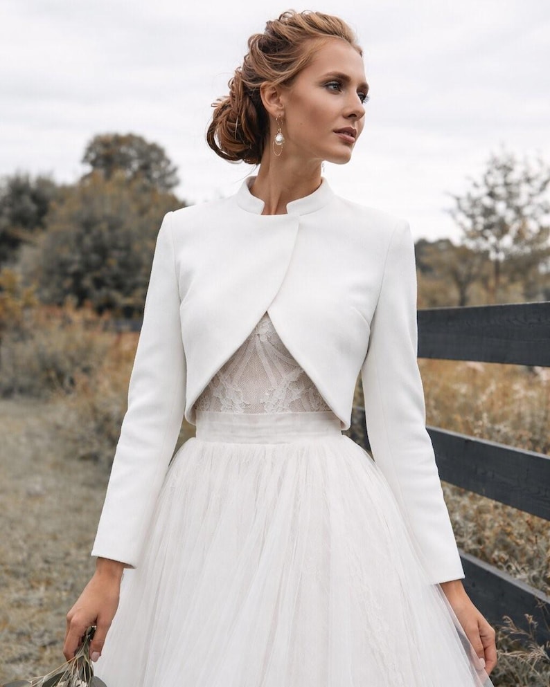 May include: A white bridal bolero jacket with long sleeves and a high collar, paired with a white tulle skirt. The jacket is tailored and covers the shoulders, creating an elegant silhouette. The background features a blurred outdoor scene.