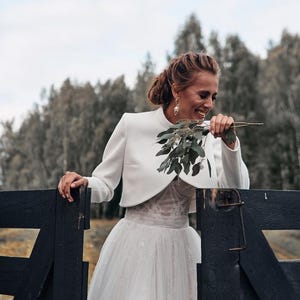 May include: A woman in a white wedding dress and bolero jacket smiles while holding greenery. The dress features a tulle skirt and lace bodice. She leans against a dark wooden fence, with a forest in the background.