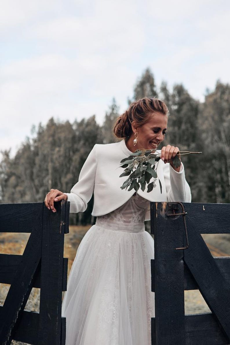 May include: A woman in a white wedding dress and bolero jacket smiles while holding greenery. The dress features a tulle skirt and lace bodice. She leans against a dark wooden fence, with a forest in the background.