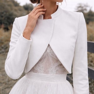 May include: A white bridal jacket with a high collar and long sleeves. The jacket is worn over a white lace bodice and a tulle skirt. The jacket is a classic and elegant addition to a wedding dress.