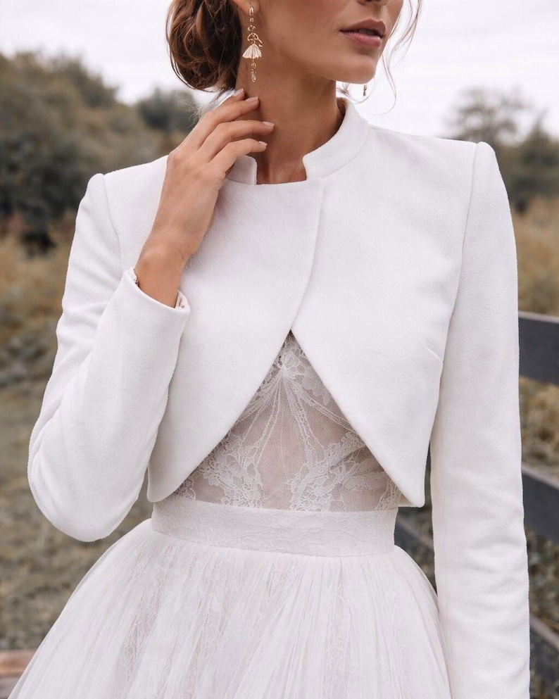 May include: A white bridal jacket with a high collar and long sleeves. The jacket is worn over a white lace bodice and a tulle skirt. The jacket is a classic and elegant addition to a wedding dress.