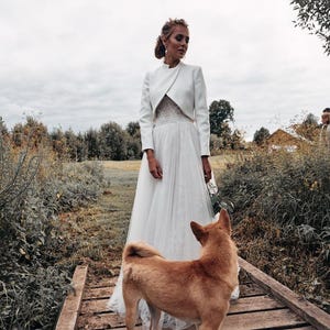 May include: A bride in a white wedding dress and jacket stands on a wooden bridge, with a brown dog looking towards her. The scene is set outdoors with greenery and a cloudy sky.