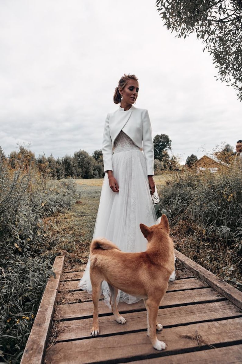 May include: A bride in a white wedding dress and jacket stands on a wooden bridge, with a brown dog looking towards her. The scene is set outdoors with greenery and a cloudy sky.