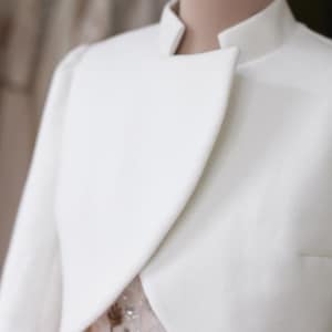 May include: A white, cropped jacket with a high collar and a unique asymmetrical design. The jacket is displayed on a mannequin, revealing a glimpse of a pink dress underneath. The style suggests formal wear, possibly bridal or evening attire.