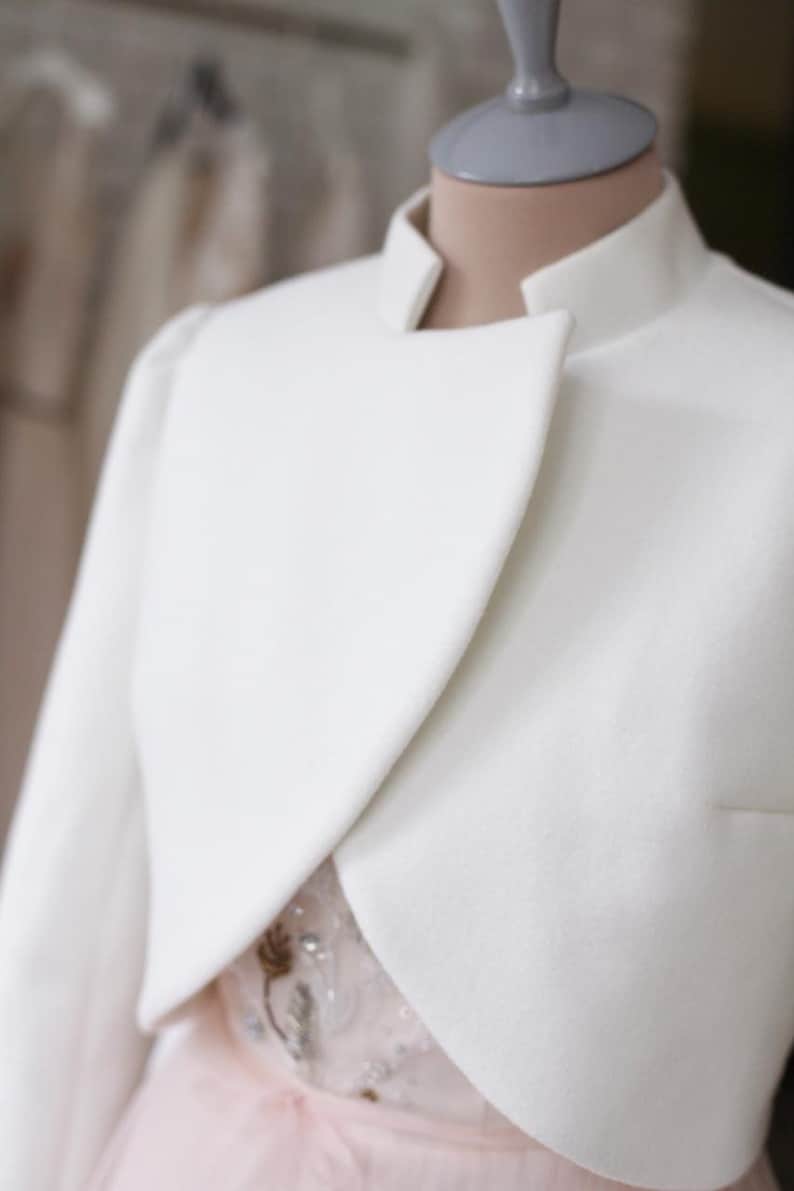 May include: A white, cropped jacket with a high collar and a unique asymmetrical design. The jacket is displayed on a mannequin, revealing a glimpse of a pink dress underneath. The style suggests formal wear, possibly bridal or evening attire.