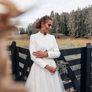 May include: A white bridal ensemble featuring a high-neck jacket and a tulle skirt. The bride is holding a small bouquet of greenery. The background includes a black wooden fence and a field with trees.