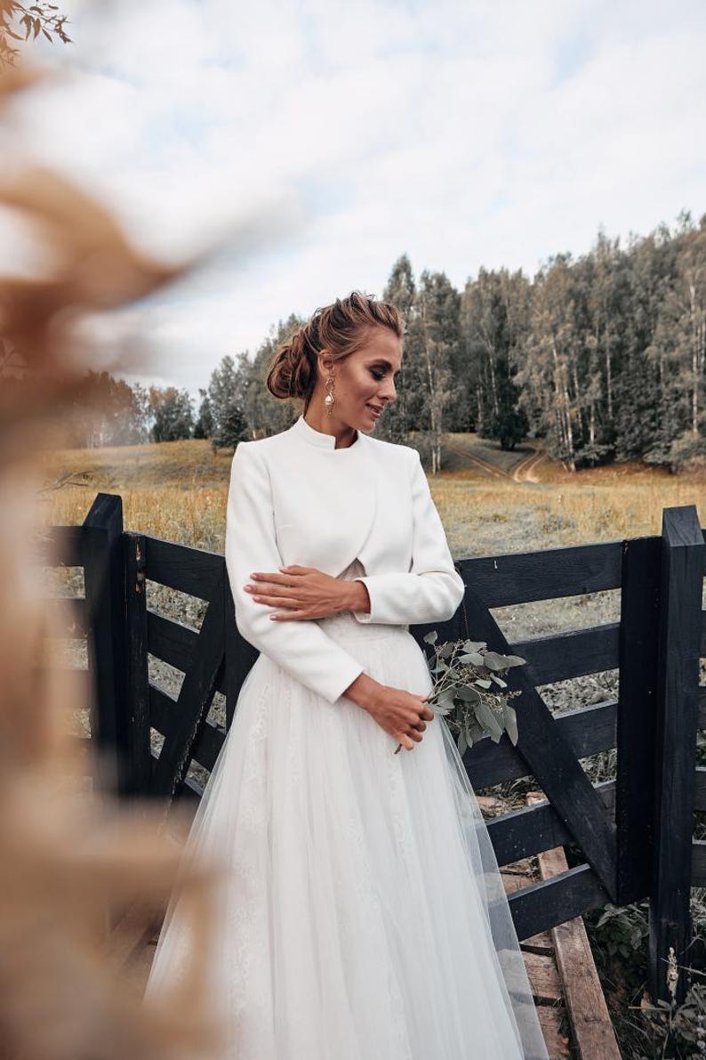 May include: A white bridal ensemble featuring a high-neck jacket and a tulle skirt. The bride is holding a small bouquet of greenery. The background includes a black wooden fence and a field with trees.