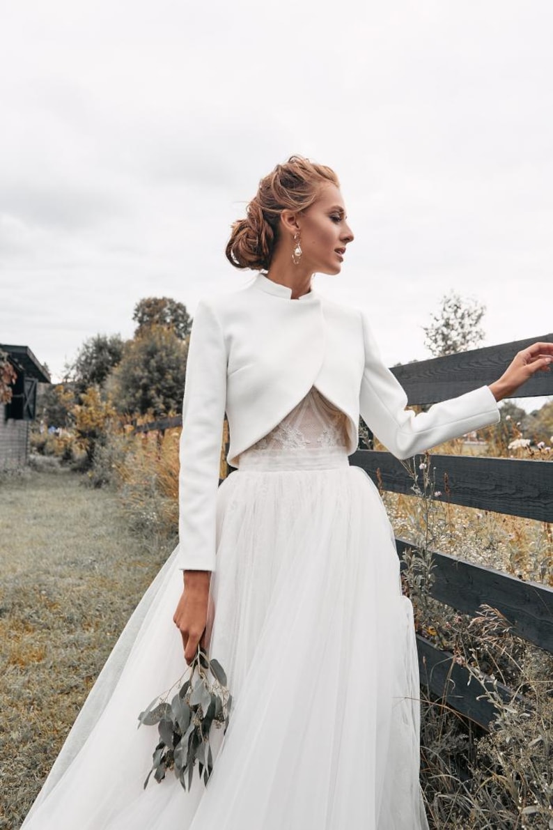 May include: A white bridal gown featuring a long, flowing tulle skirt and a cropped, long-sleeved jacket. The jacket has a high collar and is paired with a lace bodice. The bride is holding a small bouquet of greenery.