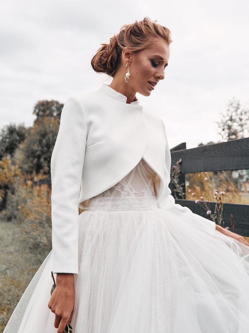 May include: A white bridal jacket with long sleeves and a high collar, paired with a white tulle skirt. The jacket is tailored and elegant, creating a sophisticated look. The outfit is perfect for a wedding or special occasion.
