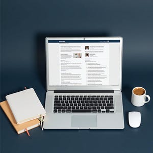 May include: A laptop displaying a webpage, with a white mouse and a white mug of coffee on a dark blue surface. Two notebooks with a pen are on the left. The laptop keyboard is visible.