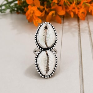 Puede incluir: Un anillo de tono plateado que presenta dos conchas de cauri blancas apiladas verticalmente. Cada concha está engastada en un borde de metal plateado con cuentas, creando una forma ovalada alargada. La banda del anillo es visible a los lados.