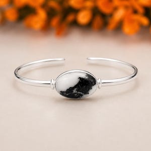 May include: A silver cuff bracelet featuring an oval-shaped white and black stone centerpiece. The bracelet has a simple, open design with a polished finish. The stone has a marbled appearance, adding a unique touch to the jewelry.
