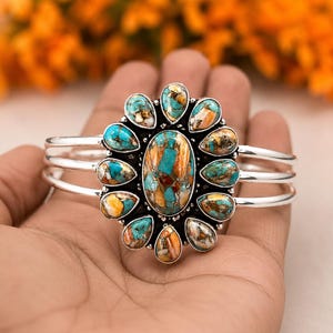 May include: A silver cuff bracelet with a large, oval turquoise and brown stone centerpiece, surrounded by smaller teardrop-shaped stones in similar colors. The bracelet has three parallel bands.