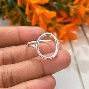 Op de afbeelding: Een ring van gepolijst zilver met een opvallend, open cirkelvormig ontwerp. De band is dun en glad en verbindt met een groot, hol ovaalvormig middendeel dat verticaal op de vinger rust. Het metaal heeft een heldere, reflecterende afwerking.