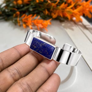 May include: A silver cuff bracelet featuring a rectangular, deep blue lapis lazuli stone. The stone is set in a polished silver frame, and the bracelet has a simple, modern design. The background includes orange flowers.