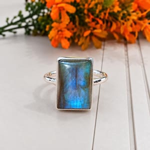 May include: A silver ring featuring a large, rectangular labradorite gemstone with a prominent blue iridescent flash. The stone is set in a smooth metal bezel, mounted on a simple, polished silver band that encircles the finger.