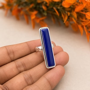 May include: A silver ring featuring a long, vertical, rectangular-shaped deep blue stone set in a smooth metal frame. The stone has a flat, polished surface and is securely mounted on a simple, polished silver band that encircles the finger.