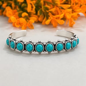 May include: A silver cuff bracelet featuring a series of oval-shaped turquoise stones with dark veining. The stones are set in individual silver bezels with a twisted rope texture and small silver bead accents between each stone setting.