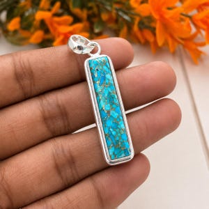 May include: A rectangular pendant featuring a vibrant turquoise stone with a mosaic-like pattern of bright blue fragments and metallic copper-coloured veins, set within a polished silver-toned metal frame with a matching silver-toned bail.