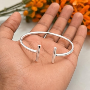 May include: A silver open bangle bracelet with two vertical bar features. The bracelet is held in a hand, with a blurred background of orange flowers.