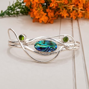 Può includere: Un bracciale rigido in argento con un intarsio centrale ovale in conchiglia di abalone dai toni blu e verdi iridescenti. La fascia metallica presenta un design fluido e vorticoso, accentuato da due piccole pietre di peridoto verde sfaccettate.