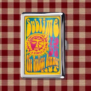 May include: A rectangular silver metal cigarette case featuring a colorful graphic print. The design includes a yellow sun with a face, blue background, and the text "SUBLIME 1995 THE ENDLESS BUMMER TOUR" in stylized yellow and pink lettering.