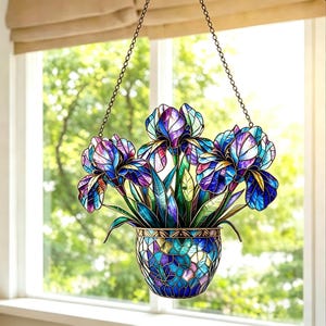 May include: A stained glass hanging planter with three iris flowers in shades of purple and blue. The flowers are in a mosaic-style pot, suspended by chains in front of a window with a blurred green background.