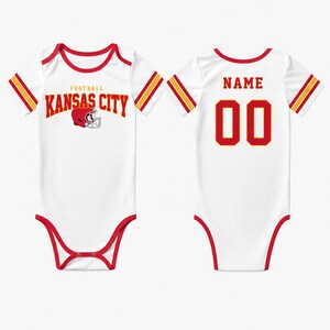May include: White baby bodysuit with red trim and accents. The front features "FOOTBALL KANSAS CITY" in red lettering with a helmet graphic. The back displays "NAME" above the number "00" in red. Sleeves have red and gold stripes.