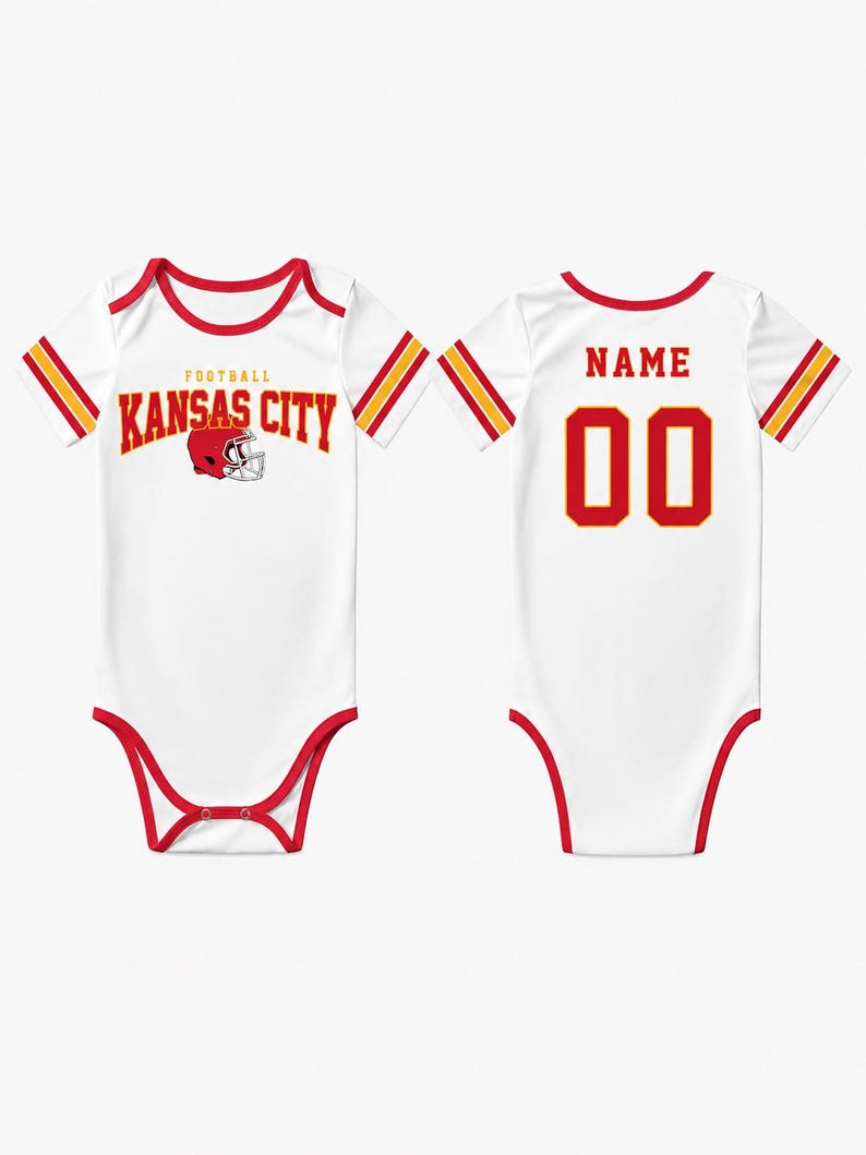 May include: White baby bodysuit with red trim and accents. The front features "FOOTBALL KANSAS CITY" in red lettering with a helmet graphic. The back displays "NAME" above the number "00" in red. Sleeves have red and gold stripes.