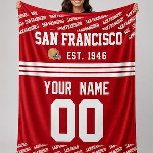 Puede incluir: Manta roja con texto y gráficos blancos. La manta presenta las palabras "SAN FRANCISCO" y "EST. 1946", junto con un gráfico de casco de fútbol. También incluye espacio para un nombre y el número "00".