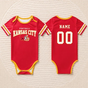 May include: Red baby bodysuit with yellow trim and white and yellow stripes on the sleeves. The front features the text "FOOTBALL KANSAS CITY" with a football helmet graphic. The back has the text "NAME" above the number "00".