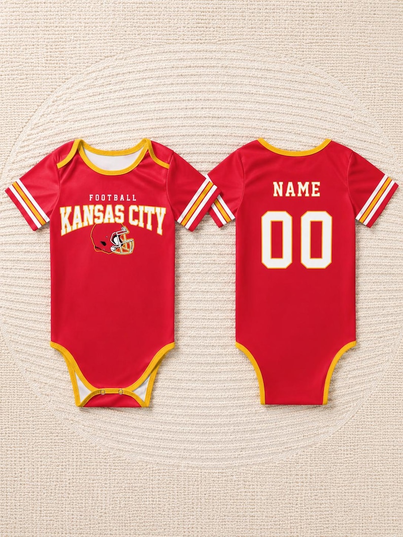 May include: Red baby bodysuit with yellow trim and white and yellow stripes on the sleeves. The front features the text "FOOTBALL KANSAS CITY" with a football helmet graphic. The back has the text "NAME" above the number "00".