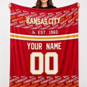 Könnte beinhalten: Rote Fleece-Decke mit dem Text "Kansas City" in Weiß und Gold sowie dem Jahr "EST. 1960". Die Decke hat ein Trikot-Design mit dem Text "YOUR NAME" und der Nummer "00" in Weiß. Die Worte "Kansas City" wiederholen sich diagonal.