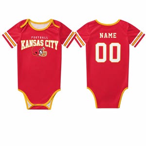 May include: Red baby bodysuit with yellow trim and white and gold stripes on the sleeves. The front features "FOOTBALL KANSAS CITY" in white text with a helmet graphic. The back has "NAME" above the number "00" in white.