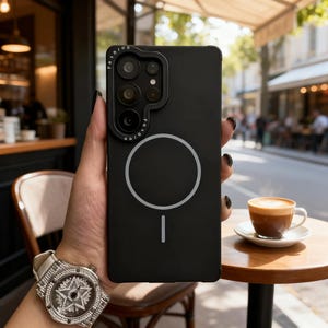 May include: A black smartphone case with a camera lens protector and a white circular design. The case has the word "Fashion" printed on the top. A cup of coffee sits on a small wooden table.