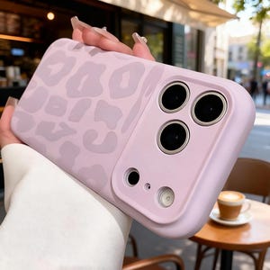 May include: A light purple phone case with a leopard print design. The case has a smooth finish and features cutouts for the phone's camera lenses and other features. The case is held in a person's hand.