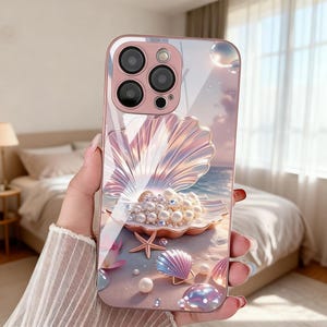 May include: A pink phone case with a seashell design, pearls, and a starfish on a beach scene. The case has a glossy finish and a raised seashell design. The camera lenses are visible at the top.