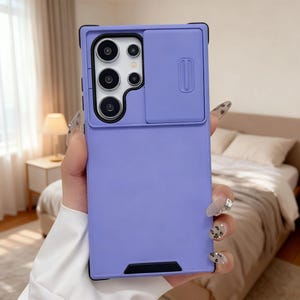 Puede incluir: Una funda de teléfono lavanda con una cubierta deslizante para la cámara. La funda tiene un marco negro y una forma rectangular. El área de la cámara tiene varios recortes circulares para las lentes. La funda se sostiene en una mano.