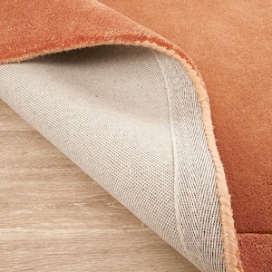 Op de afbeelding: Een terracotta-kleurig tapijt met een zacht, dichtpolig oppervlak en een zichtbare lichtbeige geweven stoffen achterkant. De hoek van het tapijt is omgeslagen om de onderkant te onthullen, met de gestikte randafwerking en de textuur van het materiaal.